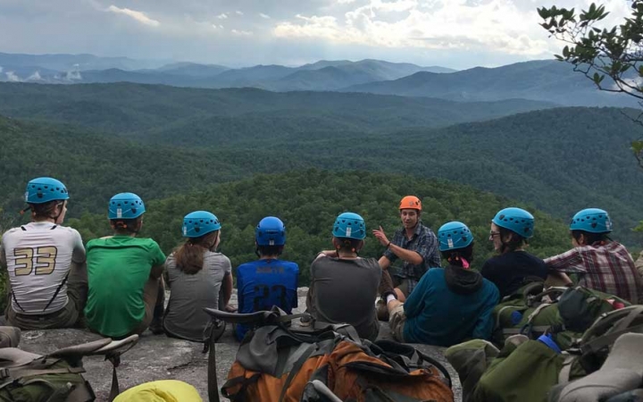 A group of students wearing helmets sit in a line and listen to an instructor speak. In the background, there is a vast and green mountainous landscape.